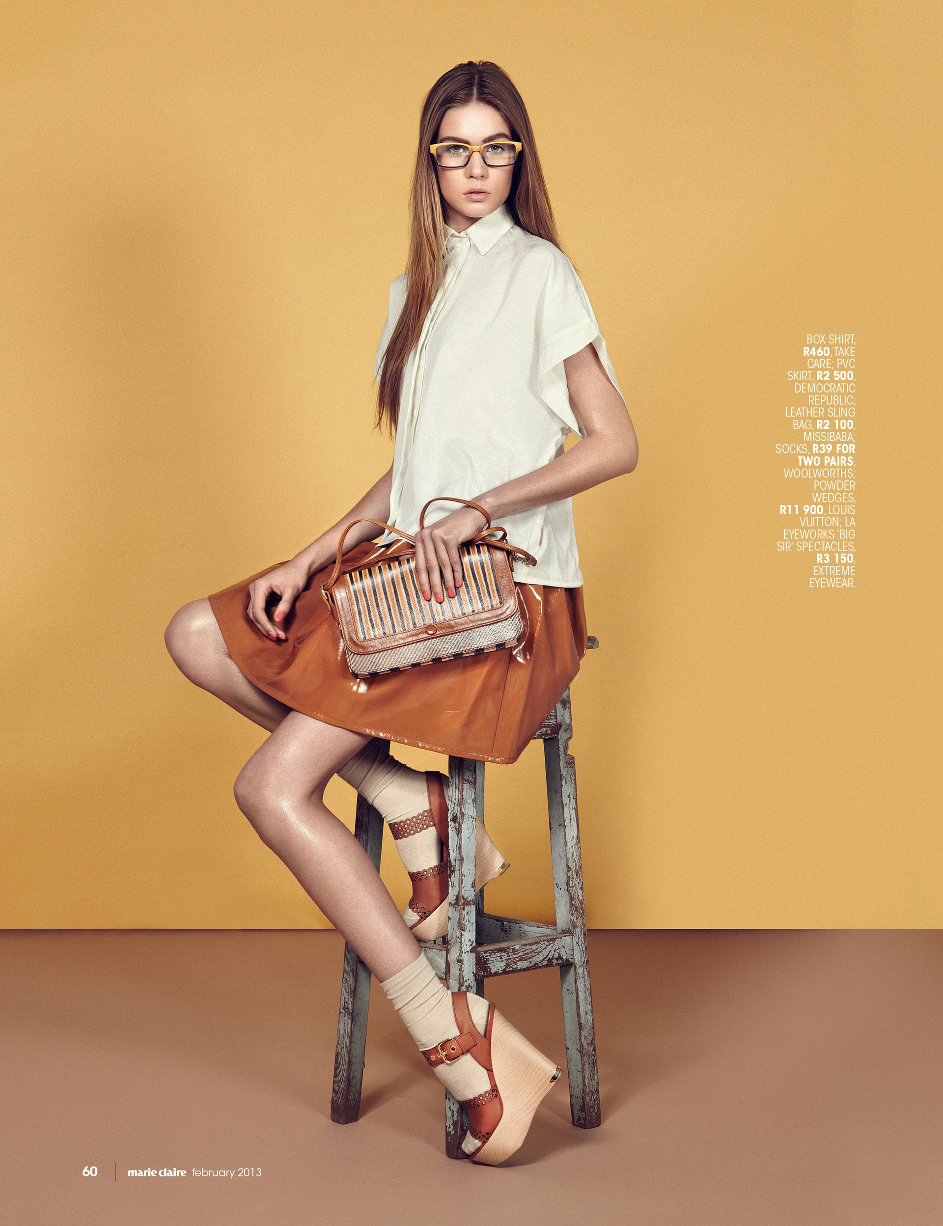 A young woman with long straight brown hair and yellow-framed glasses stands against a mustard-yellow background. She's wearing a white short-sleeved box shirt and a shiny caramel-colored PVC skirt. She carries a striped handbag with leather handles and brown details. She's perched on the edge of a weathered wooden stool, showing off mismatched socks in shades of beige, white, and rust, paired with thick wooden platform shoes with leather straps. Text on the image lists details about her outfit and accessories, and there's a marie claire February 2013 label at the bottom left.