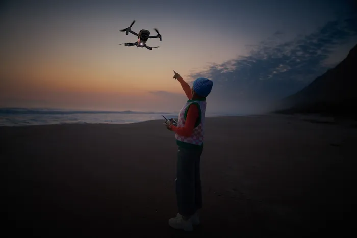 Person with remote controller pointing at a drone flying over a beach during sunset.