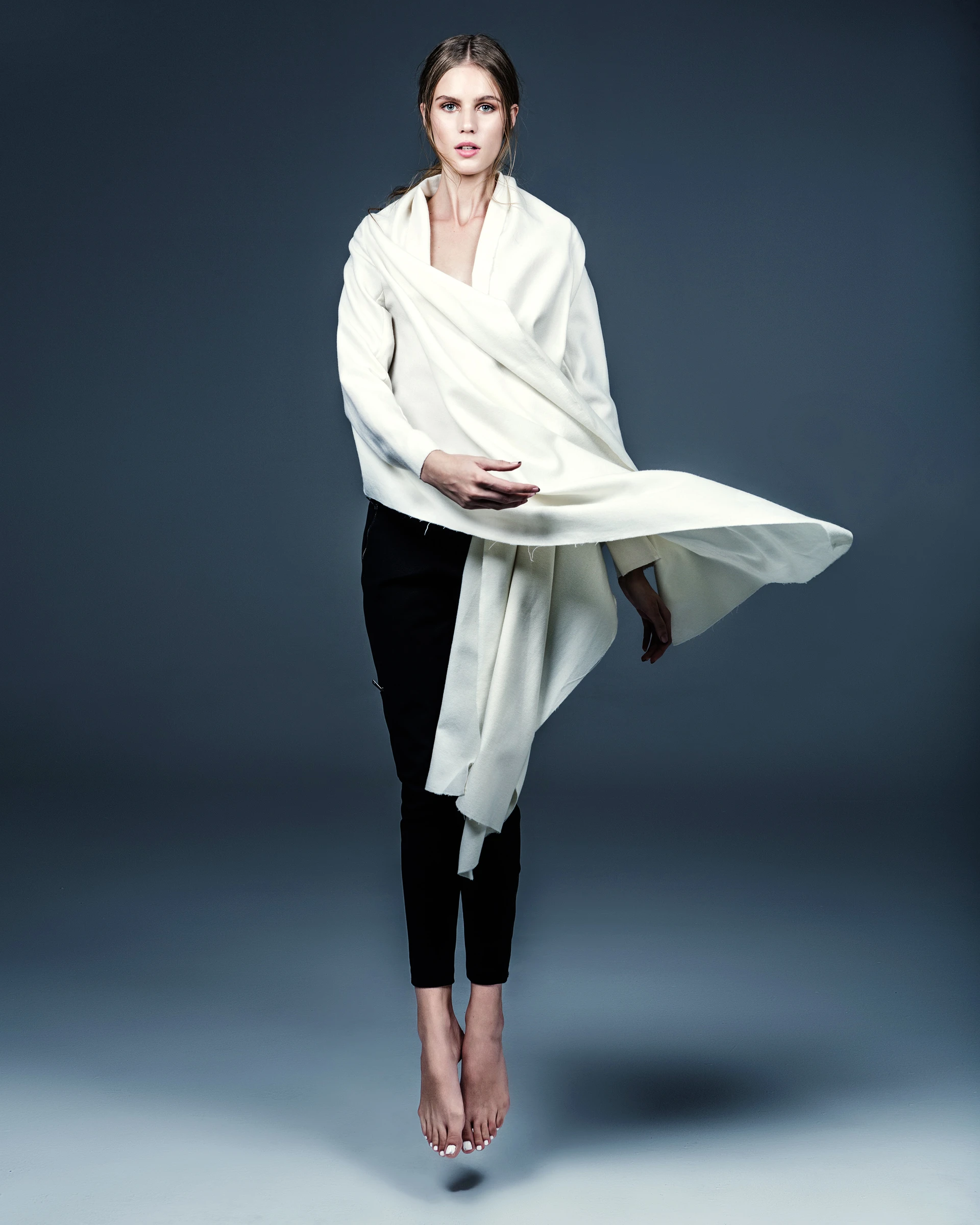 Full-length portrait of a woman in black pants and barefoot, draped in a flowing white fabric, set against a deep blue-gray background.