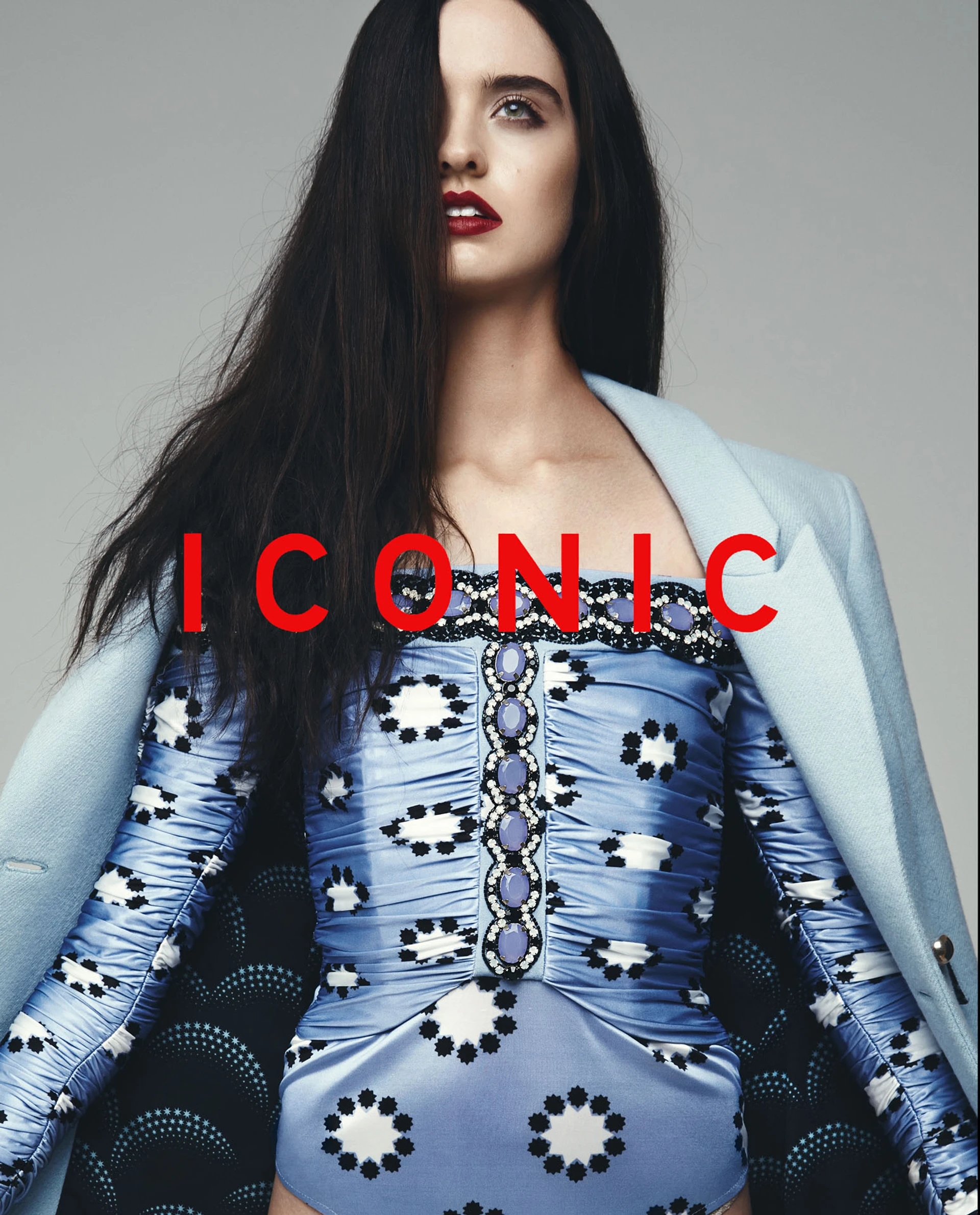 Close-up of a woman with long dark hair wearing a blue and white patterned dress, paired with a light blue coat. The word 'ICONIC' is prominently displayed across the image for Wanted Magazine.