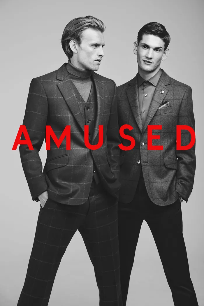 Two men in stylish dark suits, one with a slicked back hairstyle and the other with short hair, posing against a gray background. The word 'AMUSED' is overlaid in bold red letters for Wanted Magazine.